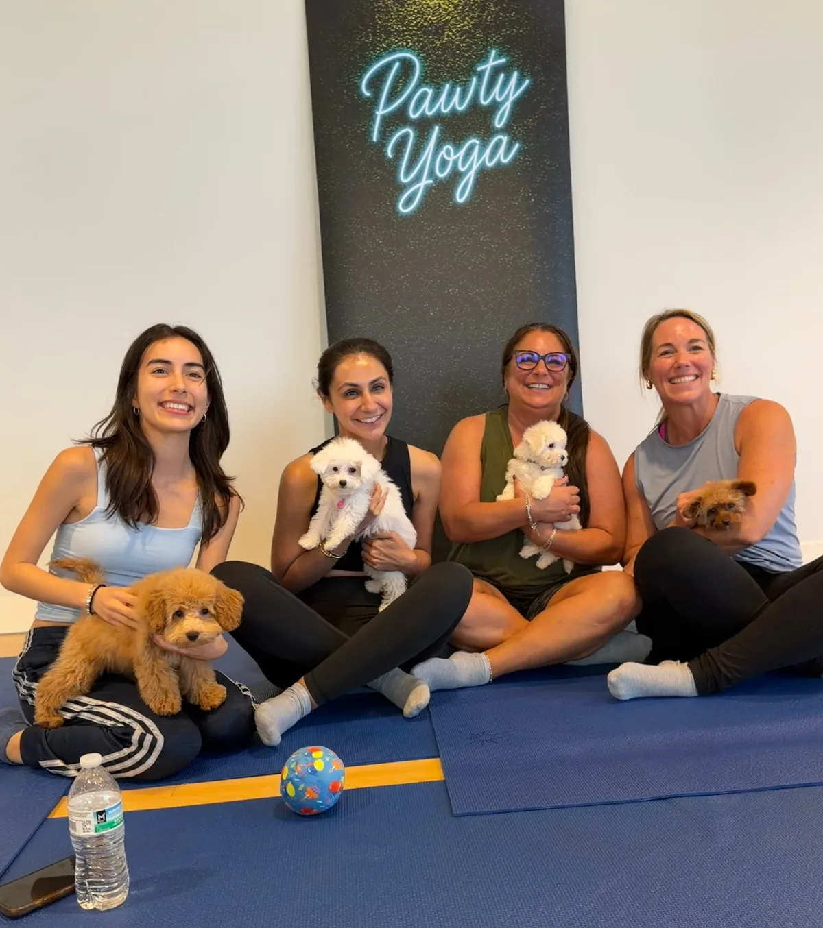Three friends holding puppies under the Pawty Yoga neon sign at a Houston cuddle session