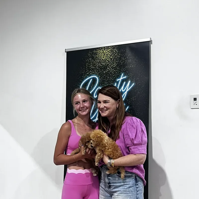 Two friends holding a puppy in front of the Pawty Yoga sign, a perfect Galentine's photo